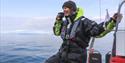 En smilende guide om bord en RIB båt på en fjord som tar seg en kopp med varm drikke
