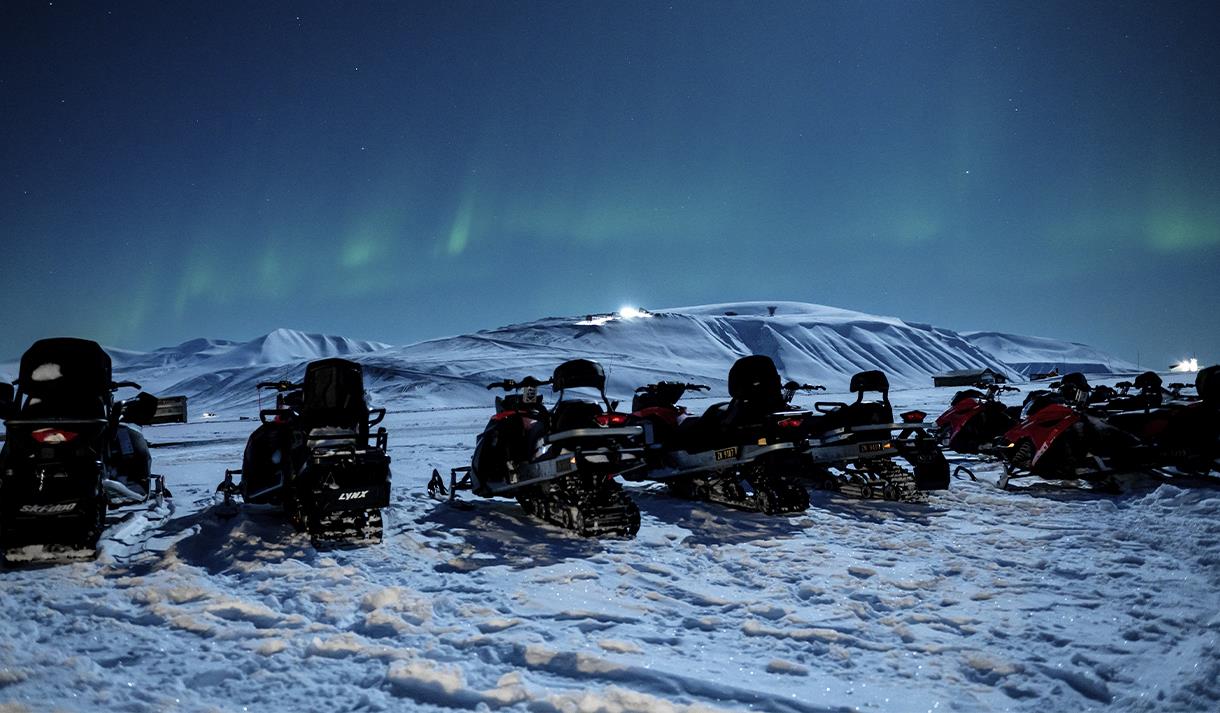 Snøscootere parkert på en rekke med svake nordlys og snødekte fjell i bakgrunnen