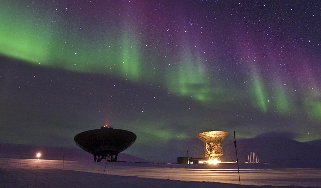 To radarantenner som peker opp mot en stjerneklar himmel med grønt og lilla nordlys dansende i bakgrunnen.