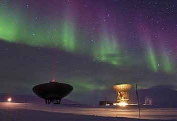 To radarantenner som peker opp mot en stjerneklar himmel med grønt og lilla nordlys dansende i bakgrunnen.