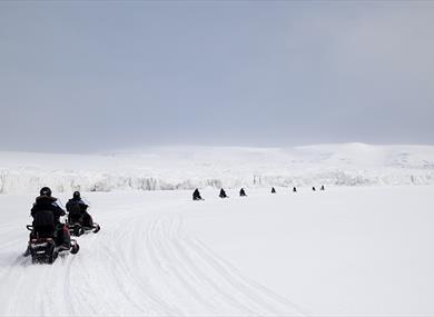 Guests in the foreground and background driving snowmobiles in a line through a snow-covered landscape, with a glacier and mountains in the background