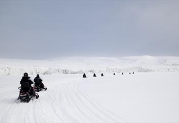 Guests in the foreground and background driving snowmobiles in a line through a snow-covered landscape, with a glacier and mountains in the background
