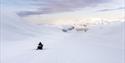 En person som kjører snøscooter på en snødekt isbre med snødekte fjell og en lett skyet himmel i bakgrunnen