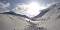 To personer som går opp et fjell langs en snødekt sti i et snøfylt fjellandskap med solskinn og en lett skyet himmel i bakgrunnen