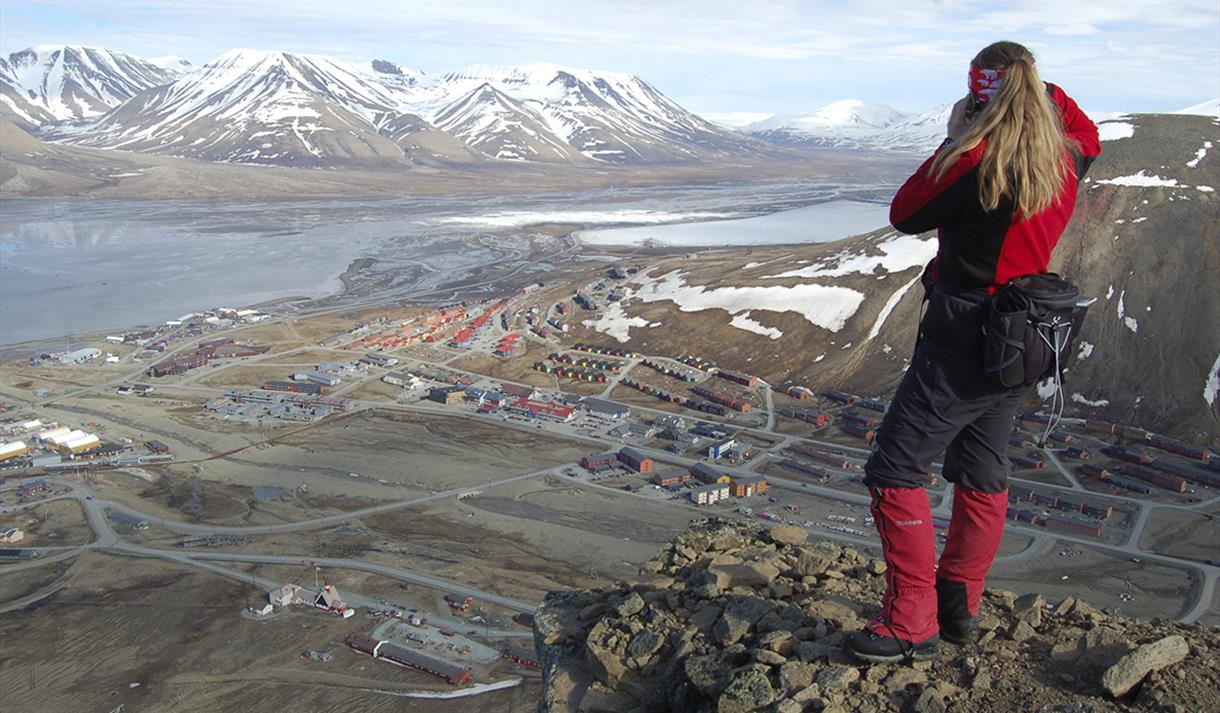En person som står på toppen av Platåfjellet og ser ned mot bygningene i Longyearbyen i bakgrunnen