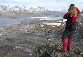 En person som står på toppen av Platåfjellet og ser ned mot bygningene i Longyearbyen i bakgrunnen