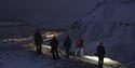A group of guests with headlamps hiking in dark surroundings, with lights from Longyearbyen in the background