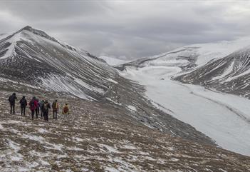 En guide og en gruppe med gjester som går ned en fjellside i forgrunnen med lett snødekte fjell og en isbre i bakgrunnen