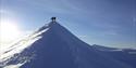 To personer på toppen av en tilsnødd fjellrygg, med snødekte fjelllandskap og en blå himmel i bakgrunnen
