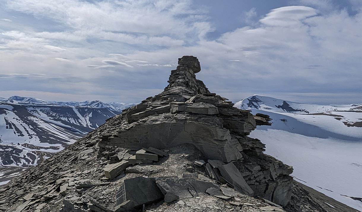 A mountain ridge with the rock formation known as Trolllsteinen, with lightly snow-covered mountains and a cloudy sky in the background