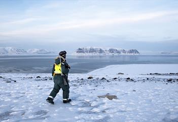A guide in snowmobile equipment carrying a rifle while walking through a snow-covered rocky field with a fjord and mountains in the background