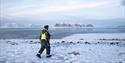 A guide in snowmobile equipment carrying a rifle while walking through a snow-covered rocky field with a fjord and mountains in the background