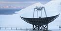 A large radar dish pointed up towards the sky with a snow-covered mountainous landscape and a fjord in the background