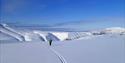 En skikjører som glir ned et snødekt fjell i forgrunnen med videre fjellkjeder og en blå himmel i bakgrunnen