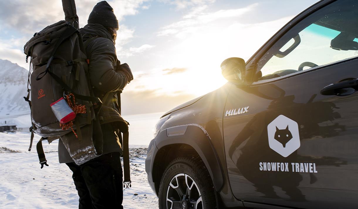 En person med en sekk og rifle på ryggen som står ved siden av en bil med solskinn, en fjord og et fjell i bakgrunnen