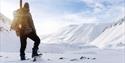 En person med en sekk og rifle på ryggen med et snødekt fjellandskap i bakgrunnen
