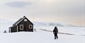 En person i snøscooterdress med en ryggsekk og rifle på ryggen som går mot en liten hytte omringet av et snødekt landskap
