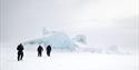 Three persons in snowmobile suits walking on snow-covered sea ice towards blue ice bergs frozen into the sea ice