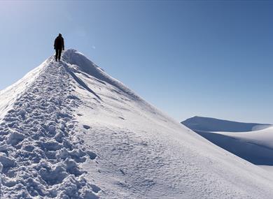 A person hiking along a mountain ridge in a snowy landscape with a clear blue sky and snowy mountains in the background