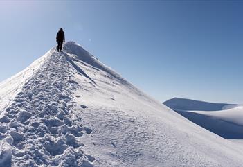 En person som går langs en fjellrygg i et snødekt landskap med en skyfri blå himmel og snødekte fjell i bakgrunnen