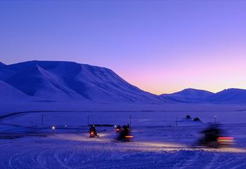 Persons on snowmobiles heading out on a trip with a mountainous landscape covered in blue light in the background