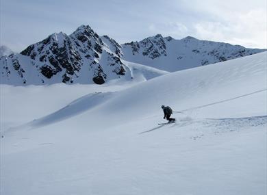 A skier skiing turns down a snow-covered mountain side in the foreground, with a range of mountains in the background