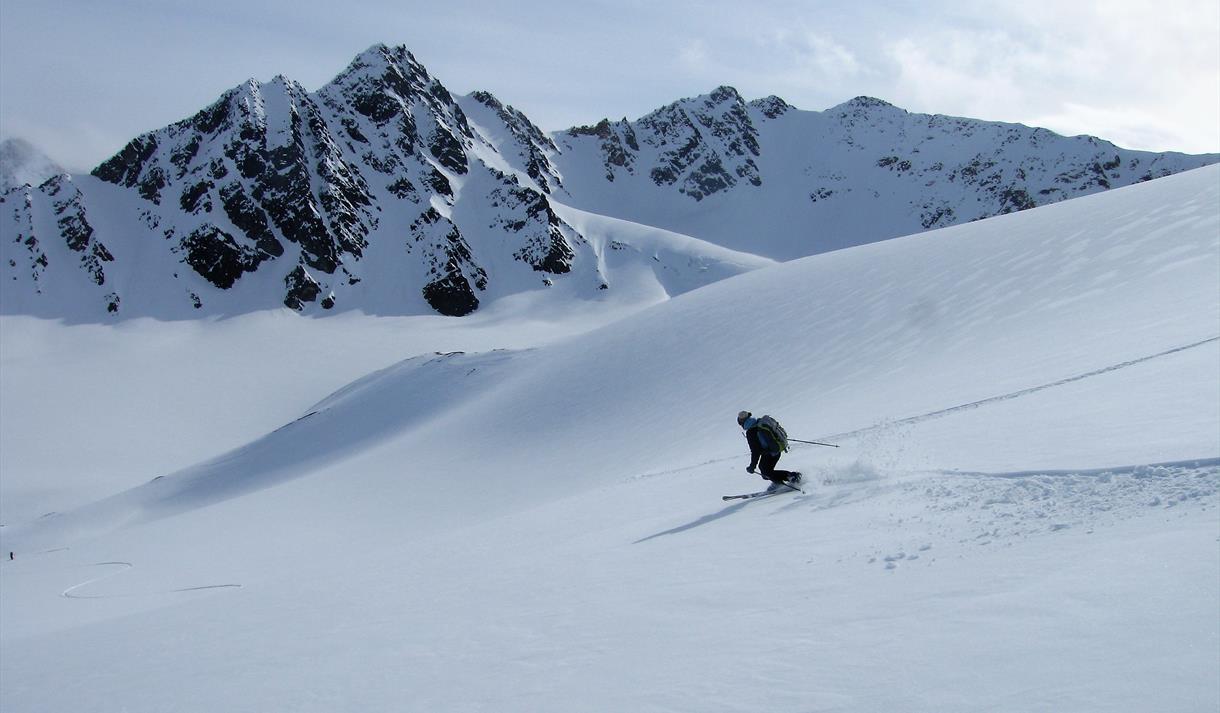 En skikjører som svinger på ski ned en snødekt fjellside i forgrunnen, med en fjellrekke i bakgrunnen