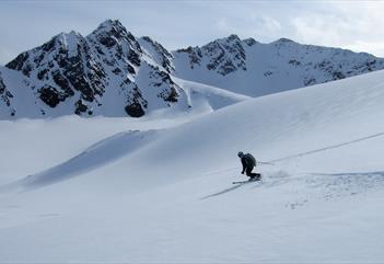 En skikjører som svinger på ski ned en snødekt fjellside i forgrunnen, med en fjellrekke i bakgrunnen