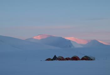 A tent camp in a snowy and shadow-covered landscape with mountains in the background whose peaks are covered in sunlight