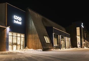 Svalbardbutikken sett utenfra i mørketiden, med lys som skinner ut fra vinduene i butikken og fra Coop Svalbard sin logo på veggen deres.