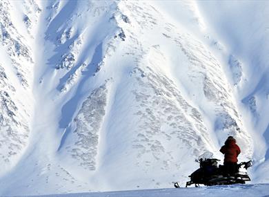 A person standing next to a snowmobile with a massive snow-covered mountainside in the background