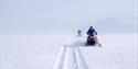 Two guests on snowmobiles driving along a track in a flat snow-covered landscape