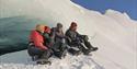 Fire personer som sitter i en snødekt bakke ved en isformasjon og speider utover et vinterlandskap