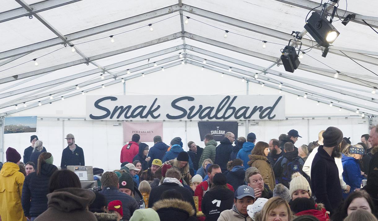 A crowd sitting in a festival tent eating, with a "Taste Svalbard" logo and a line of tables with food for sale in the background