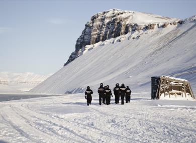 A group of guests in snowmobile equipment walking towards a building in snowy surroundings next to Villa Fredheim along Tempelfjorden, with mountains 