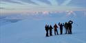 En gruppe med gjester på en snødekt fjellrygg i forgrunnen, med et vidstrakt snødekt landskap og fjerne fjell i bakgrunnen