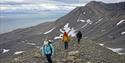 To gjester og en guide på fottur over en bakketopp i forgrunnen, med et fjell og en fjord i bakgrunnen