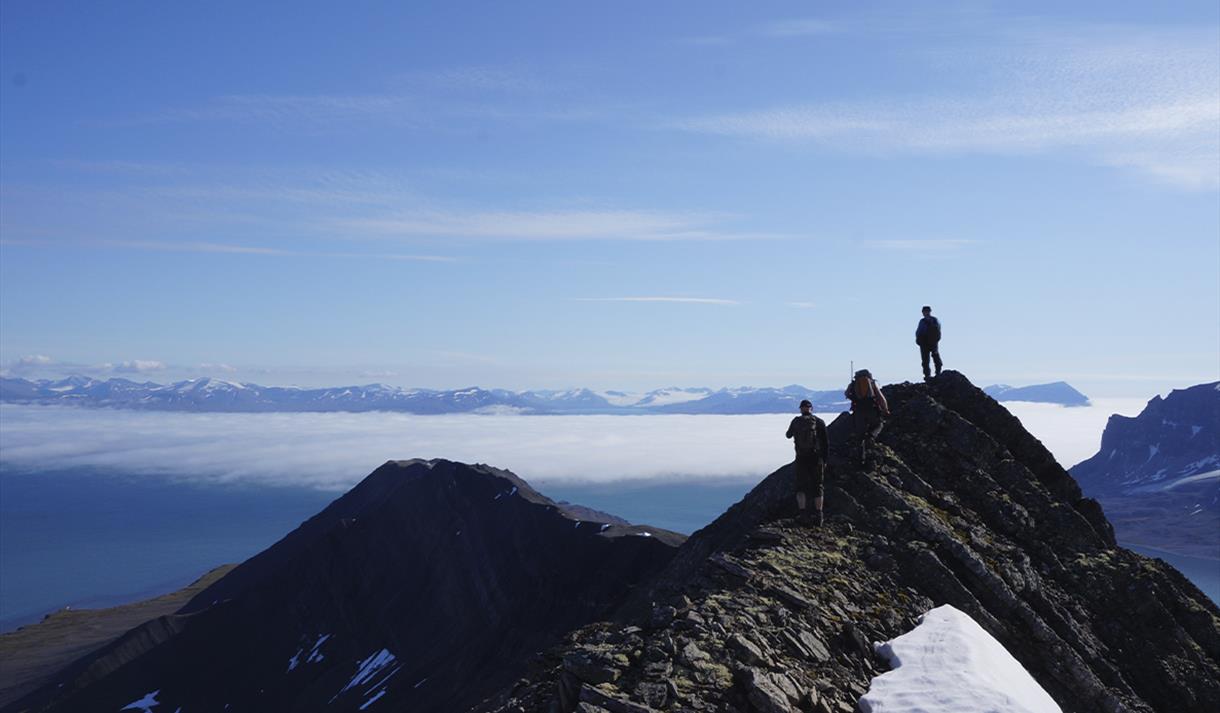 Two guests and a guide hiking along a mountain ridge in the foreground, with a vast cloud-covered fjord, distant mountains and a blue sky in the backg