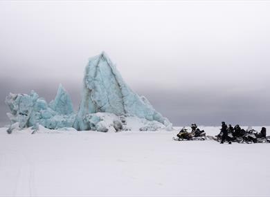 Guests and a guide on snowmobiles taking a break by an iceberg frozen in sea ice