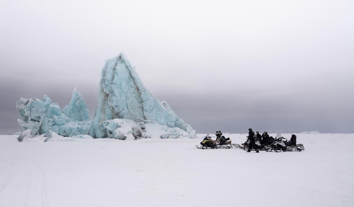 Gjester og en guide på snøscootere som tar en pause ved et isberg frosset inn i sjøis