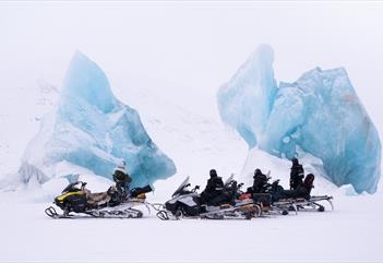 En guide og gjester med snøscootere som tar en pause ved to blå isfjell som er frosset inn i sjøis