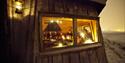 Bilde tatt fra utsiden av en hytte. På bildet ses det personer som koser seg inne i varmen med stearlinlys og bålkos