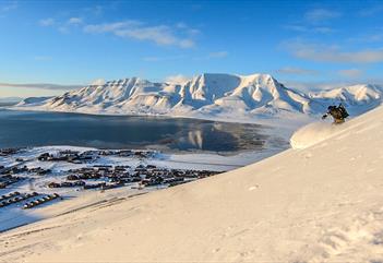 En person som står på ski ned et fjell med Longyearbyen og fjellandskap i bakgrunnen