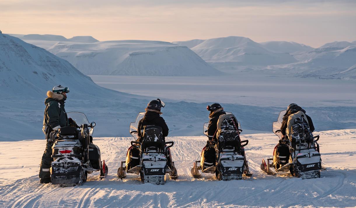 Tre gjester og en guide på snøscootere som står i ro med et snødekt landskap i bakgrunnen