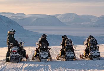 Tre gjester og en guide på snøscootere som står i ro med et snødekt landskap i bakgrunnen