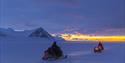 A guest and a guide driving snowmobiles at dusk. In the background there's sunlight on the horizon.