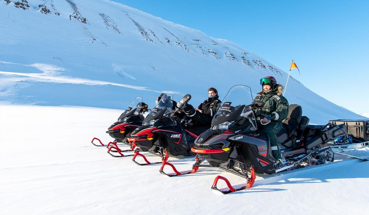 En guide og gjester på snøscootere som tar en pause på tur i et snødekt lyst landskap.
