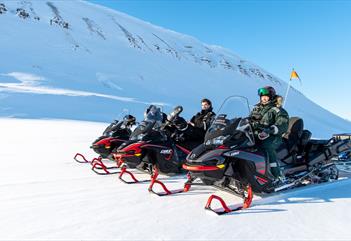 En guide og gjester på snøscootere som tar en pause på tur i et snødekt lyst landskap.