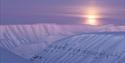 Et fjellandskap i lilla toner med en solnedgang skjult av et tynt skylag i bakgrunnen.