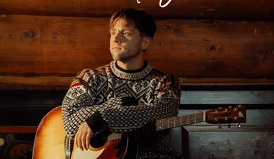 mann med strikket genser i rødt, hvitt, grått og sort med gitar